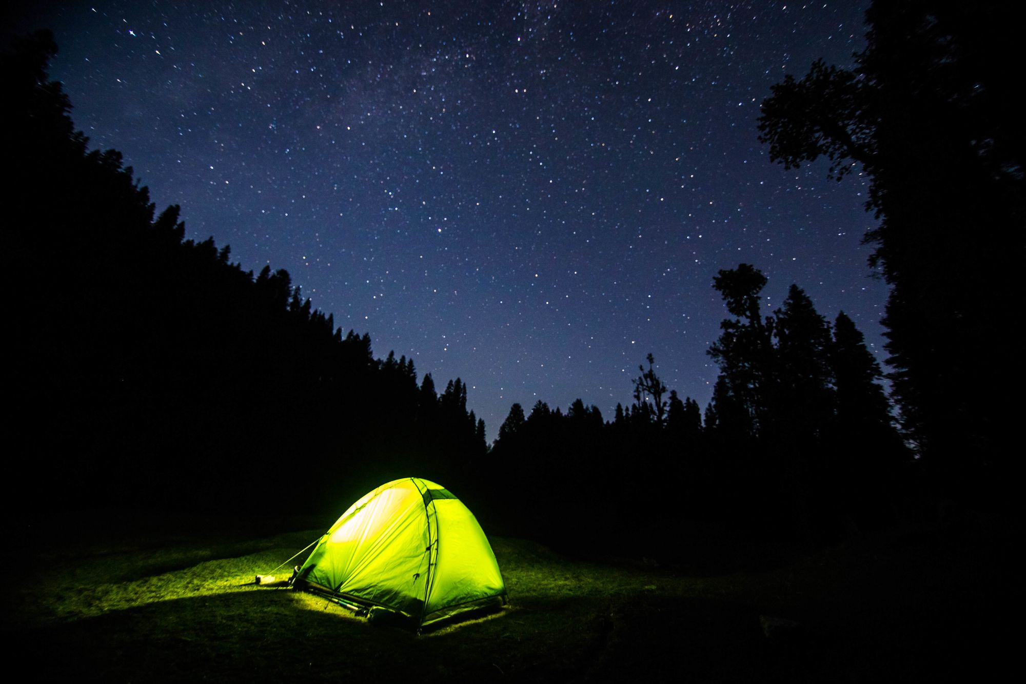 light emits from a green tent set underneath the starry sky surrounded by silhouettes of trees