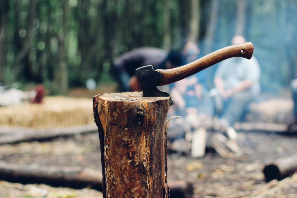 axe with a wooden handle cutting into a log with people at a campfire in the distance