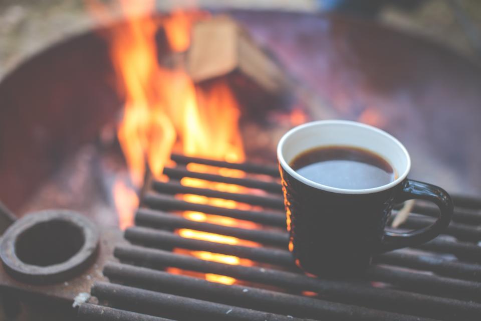 coffee in a black and white ceramic cup sits on a grill over a campfire
