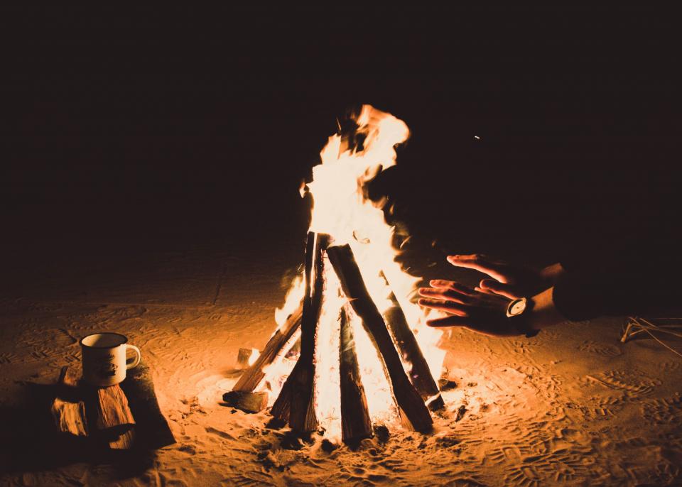 two hands held near a fire with a tall flame and sandy ground below