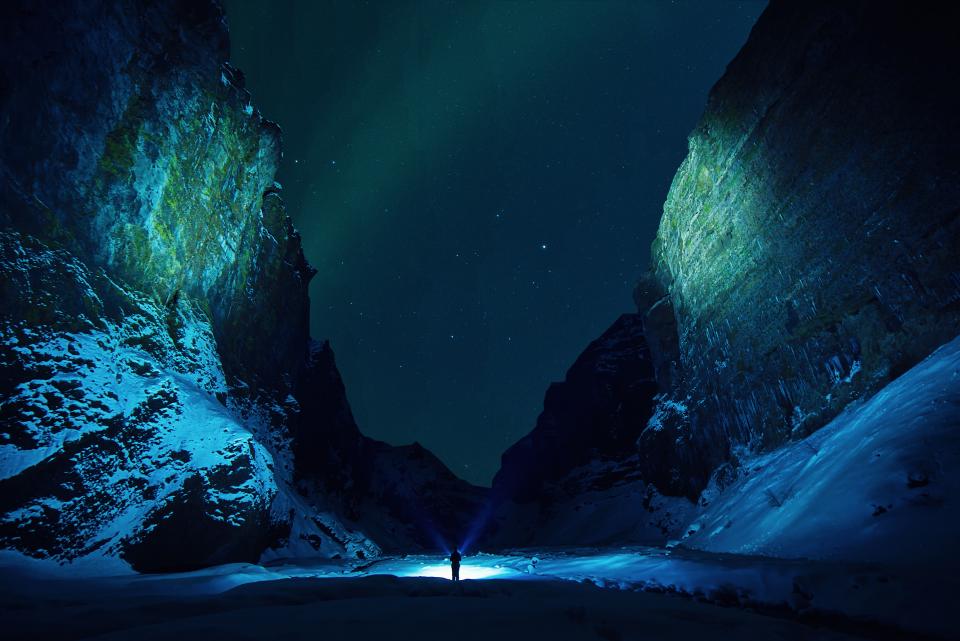 a small figure stands in between two large cave walls with auroras in the sky above