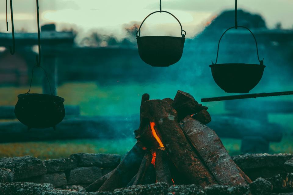 three cast-iron kettles hang over a smoldering fire pit with burning wood logs