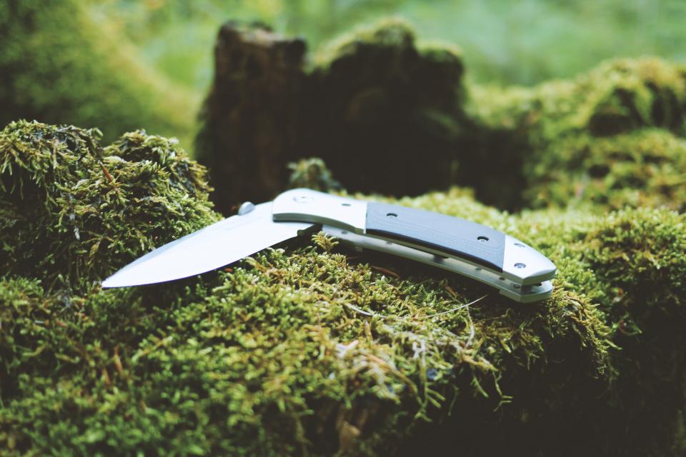 an open pocket knife with a black and silver handle sits on a moss-covered surface