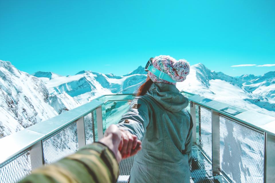 couple holds hands and cross a bridge surrounded by snowy mountains and blue sky