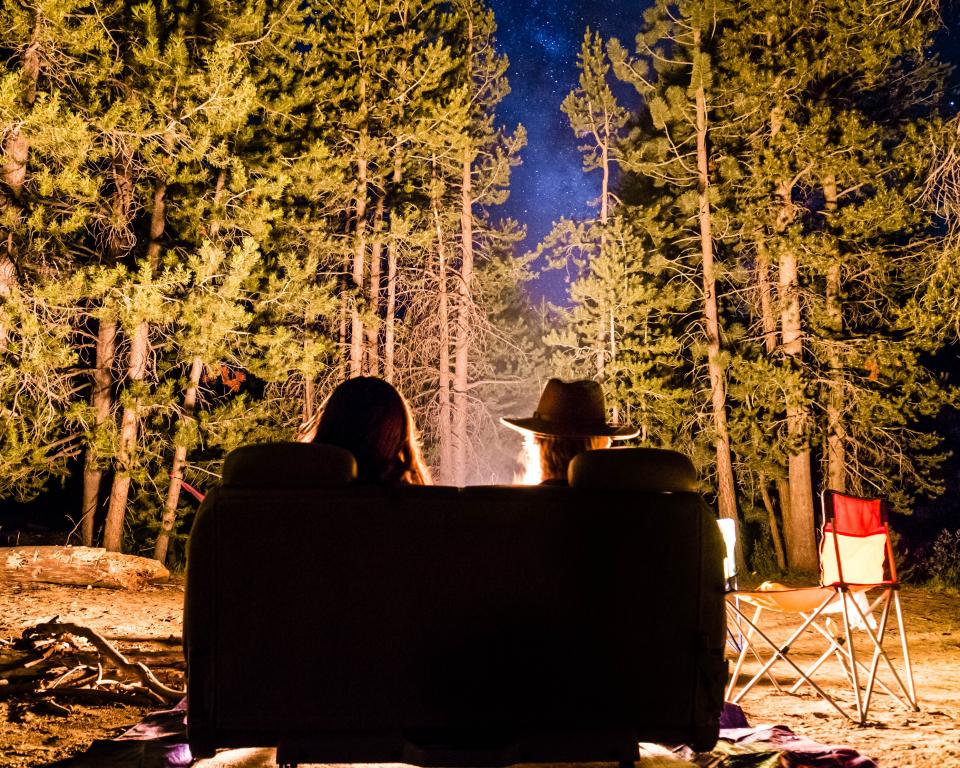 two silhouettes of people sitting by a campire surrounded by trees at night