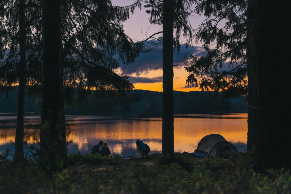 three people sit around a campfire near a lake during sunset