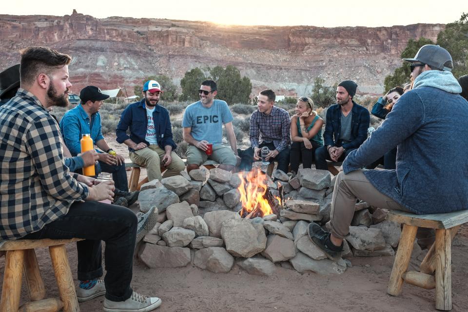 a large group of people talk and huddle around a rocky bonfire pit