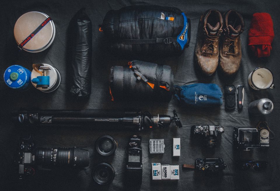 cameras, film, lenses and a tripod sit on a gray surface along with camping equipment