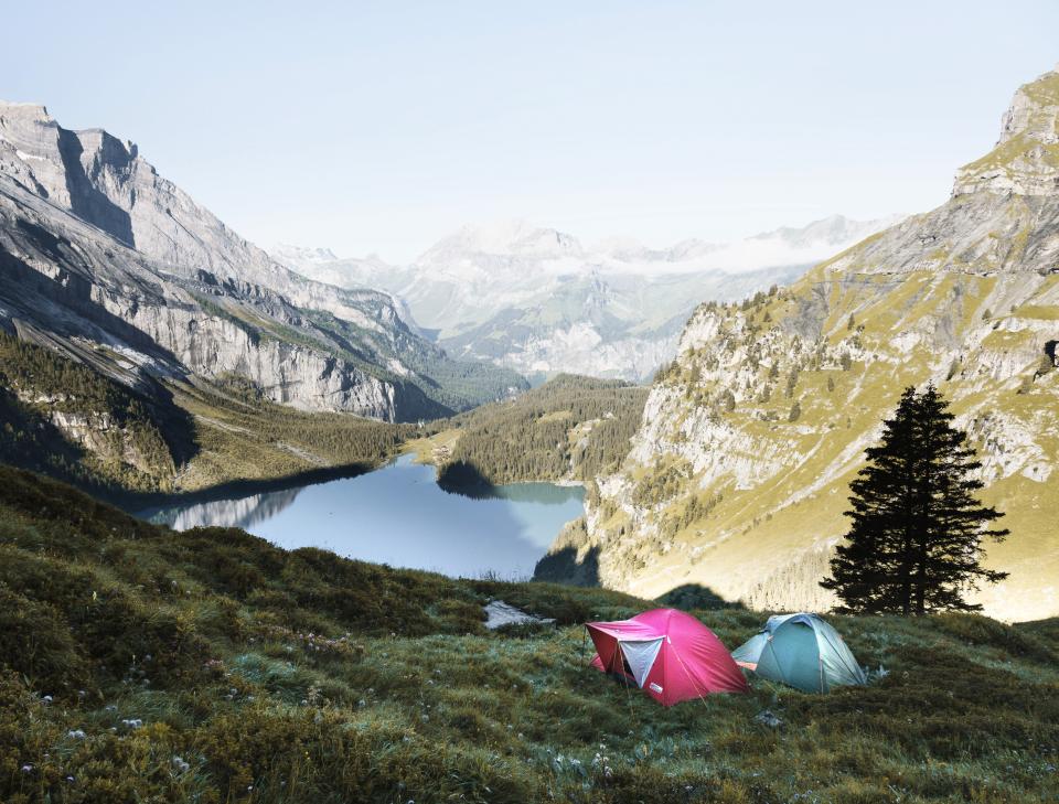 two tents sit on grass in the valley of a mountain range with water and trees in the distance