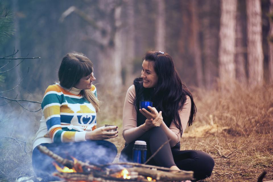 two women smile and converse over a warm drink by a campfire in the woods