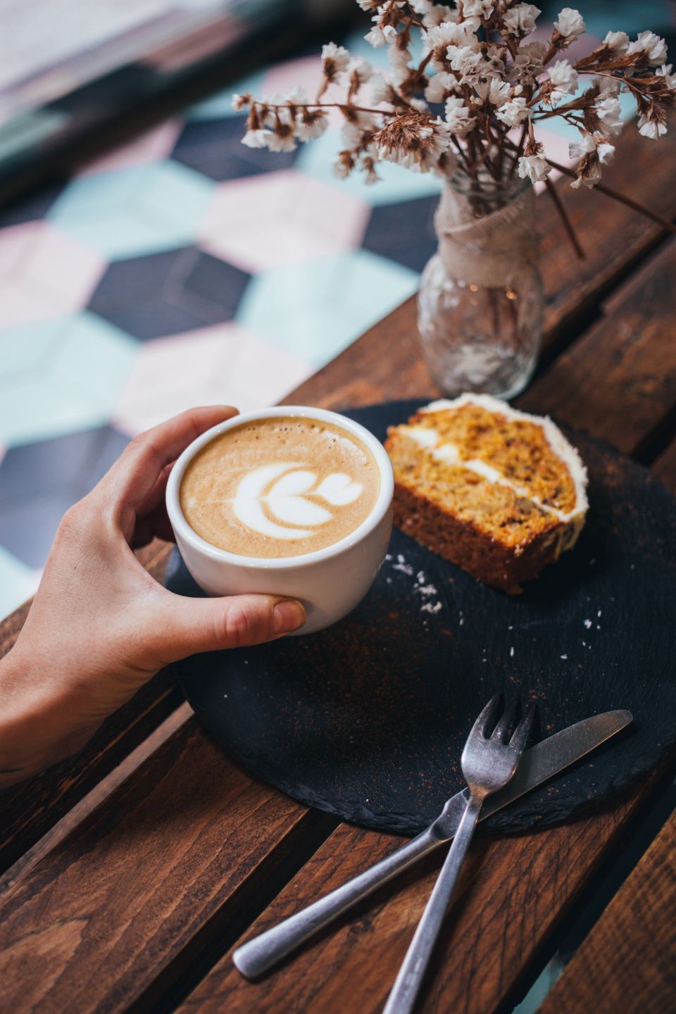 a person picks up a latte off of a platter that has utensils and a slice of carrot cake