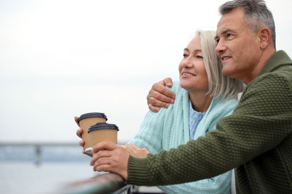 a senior couple looks out at a pier while holding to-go cups of coffee