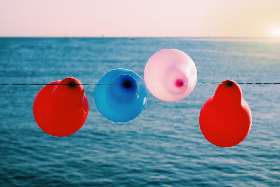 Four birthday balloons in red, blue and pink hues blow in the breeze overlooking a body of water, during sunset.