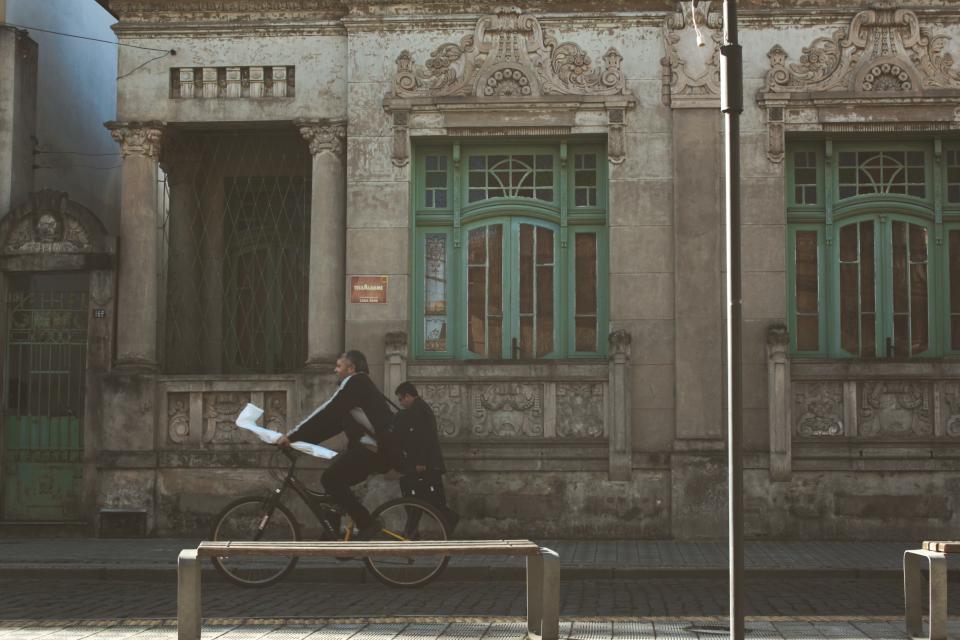 A man rides his bicycle on a cobblestone road and holds a loaf a bread and his hand.