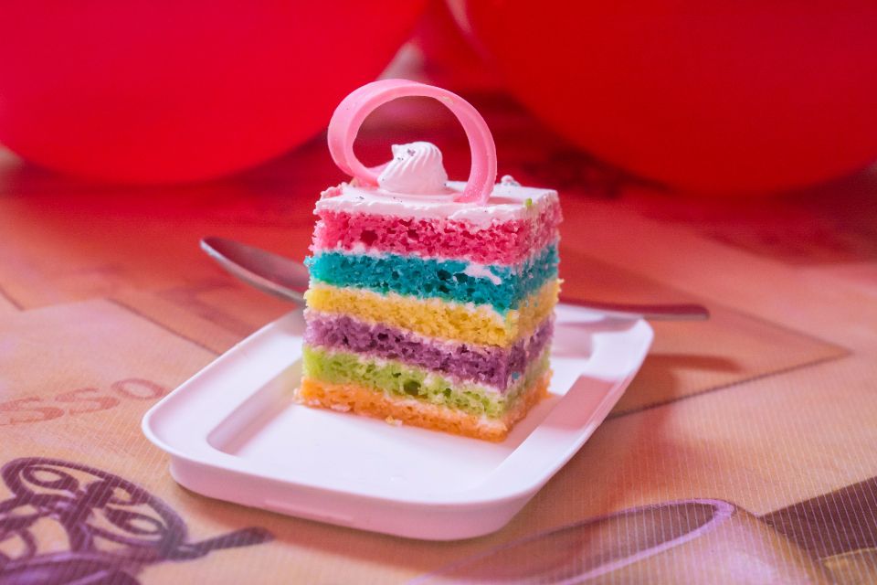 A slice of cake with rainbow-colored layers sits on a white plate with red balloons in the backgound.