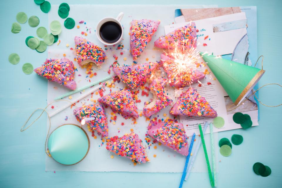Slices of a pink birthday cake with colorful, star-shaped sprinkles is surrounded by a sparkler, green party hats, confetti and a cup of coffee. 