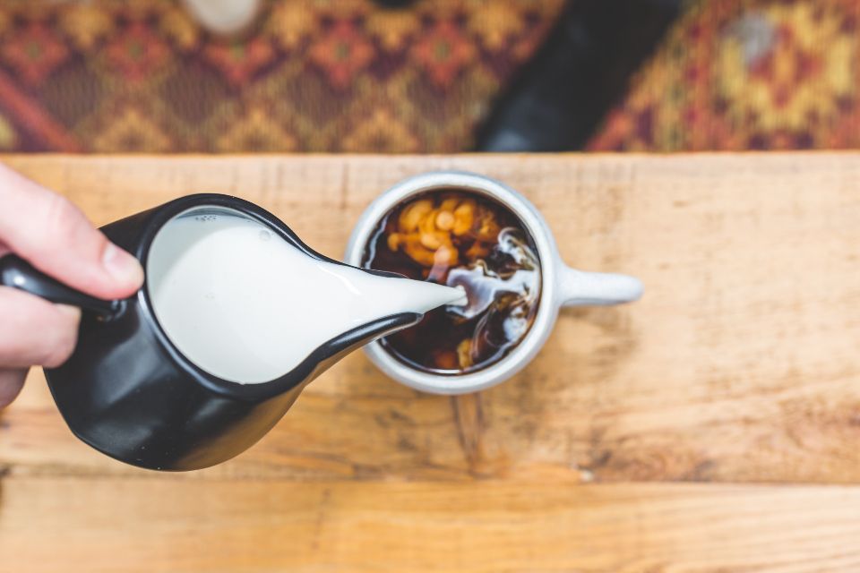 hand pours milk from a small pitcher into a cup of hot coffee