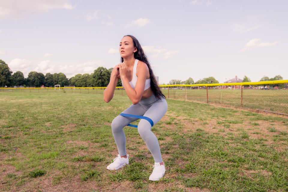 A female fitness trainer stands in a grassy field and works on squats with a resistance band.