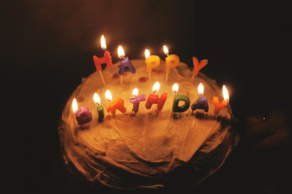 A homemade birthday cake with light-colored frosting and lit candles that spell out "happy birthday" in colorful letters.