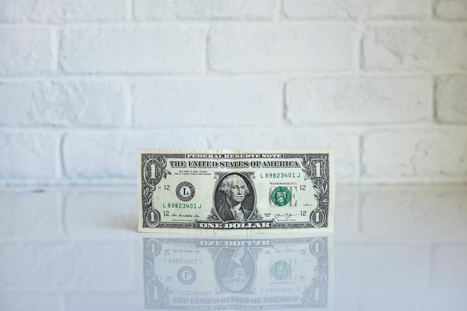 A single dollar bill sits on a white reflective surface