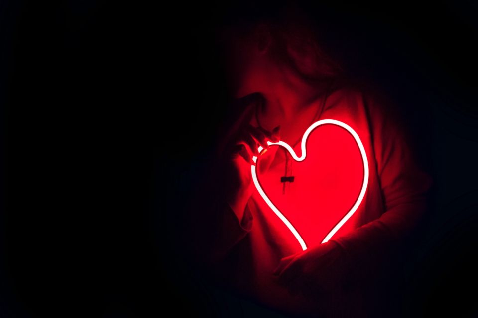 a woman holds a glowing red, heart-shaped neon sign 