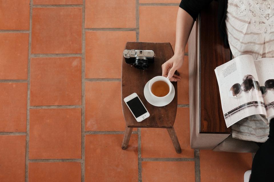 person reaches for a cup of tea that sits on a table with a phone and film camera. 