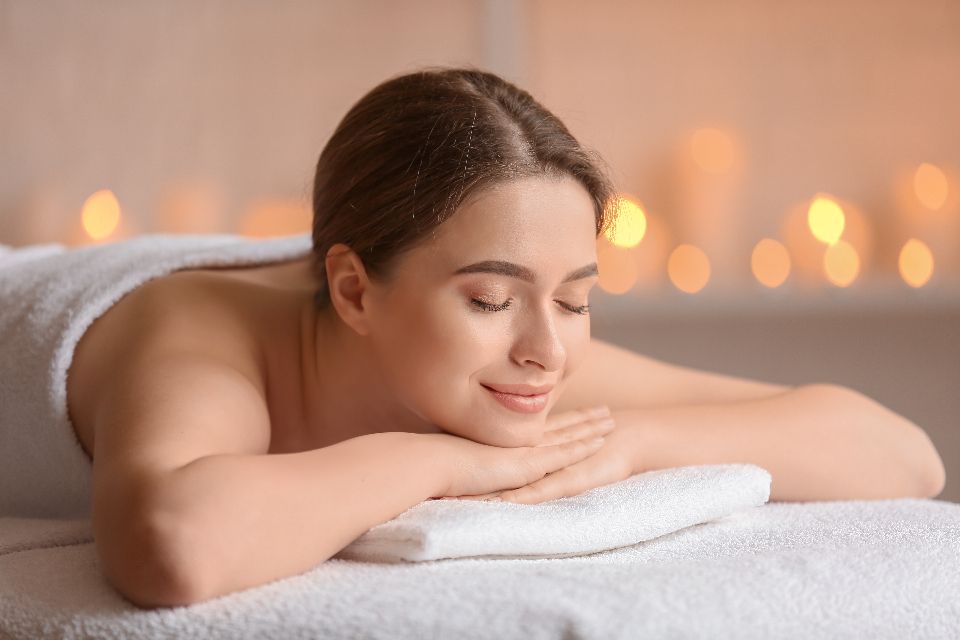 a woman wearing white towel closes her eyes and smiles while resting on a massage table, with bokeh lights in the background.