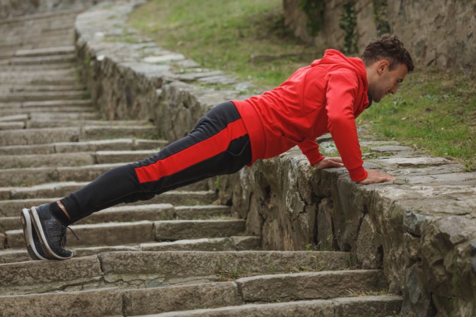 A young man in a red hoodie and black and red joggers does a push up on a stone staircase.