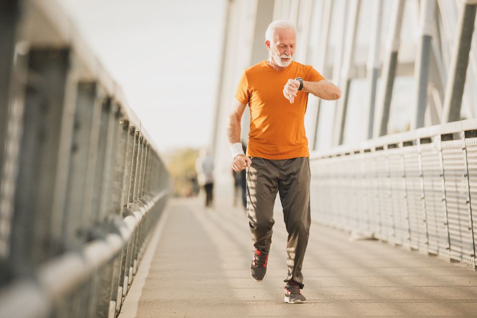 A senior man runs on a bridge, while wearing an orange shirt and looking at his watch.