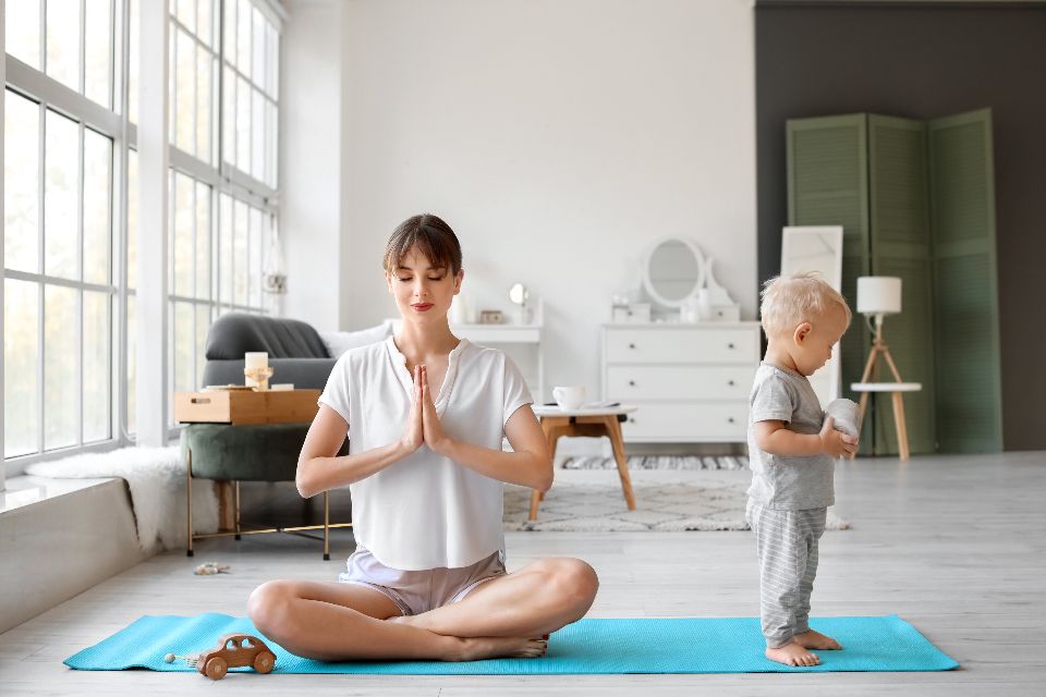 A mother sits cross-legged on a yoga mat with her toddler standing closeby.