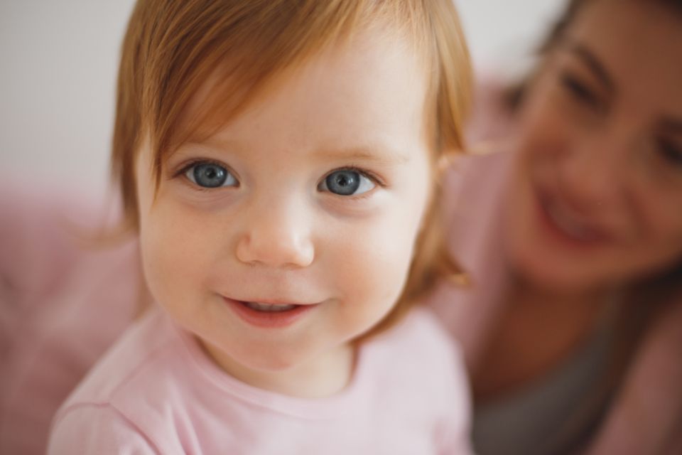 a baby girl in a pink shirt smiles sweetly with her mother in the background