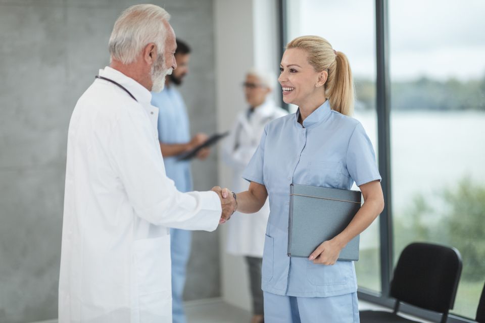 two happy healthcare professionals shake hands at a hospital with two coworkers in the distance