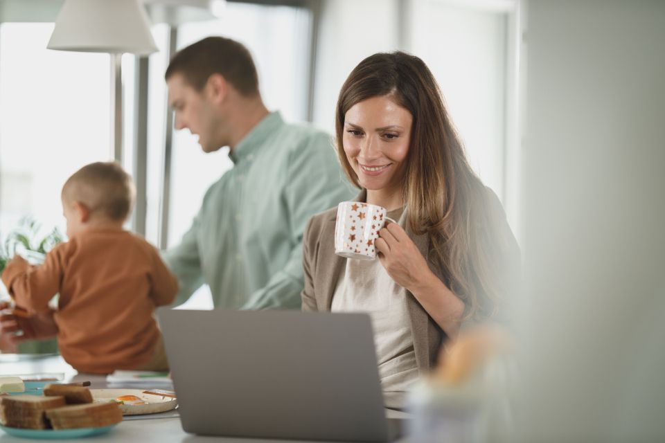 a woman smiles while working on a laptop with her husband and baby in the background 