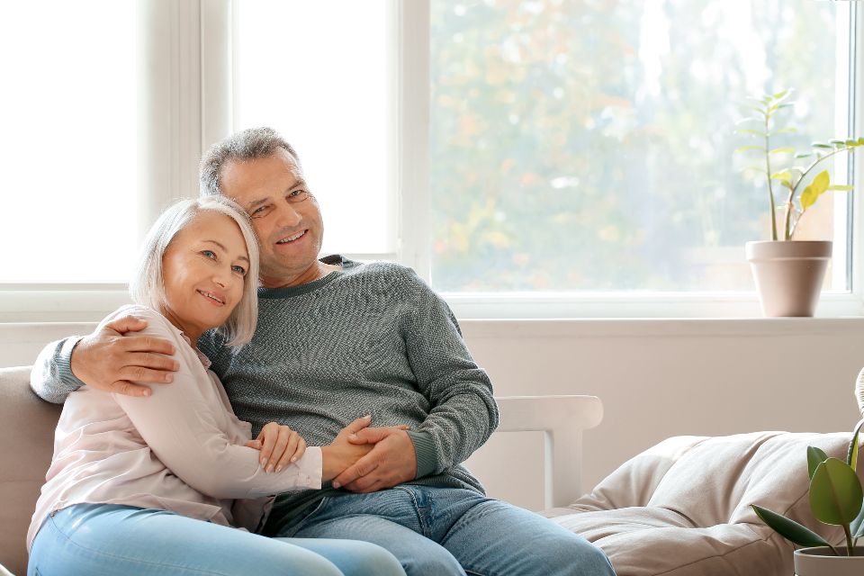 a senior couple sits on a couch near a window while hugging and smiling warmly