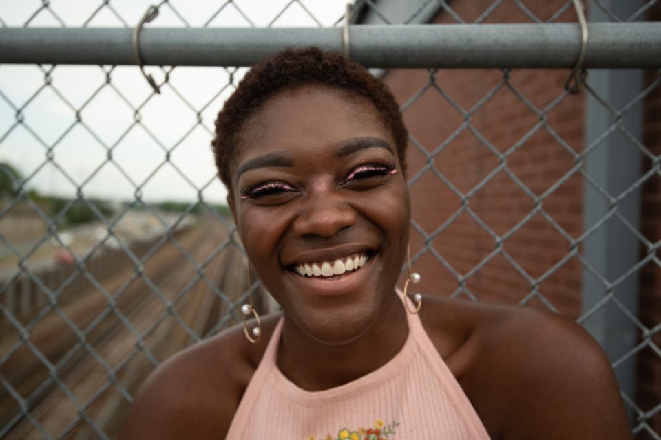 an African-american woman wearing pink eyeliner smiles and stands against a fence in an industrial landscape