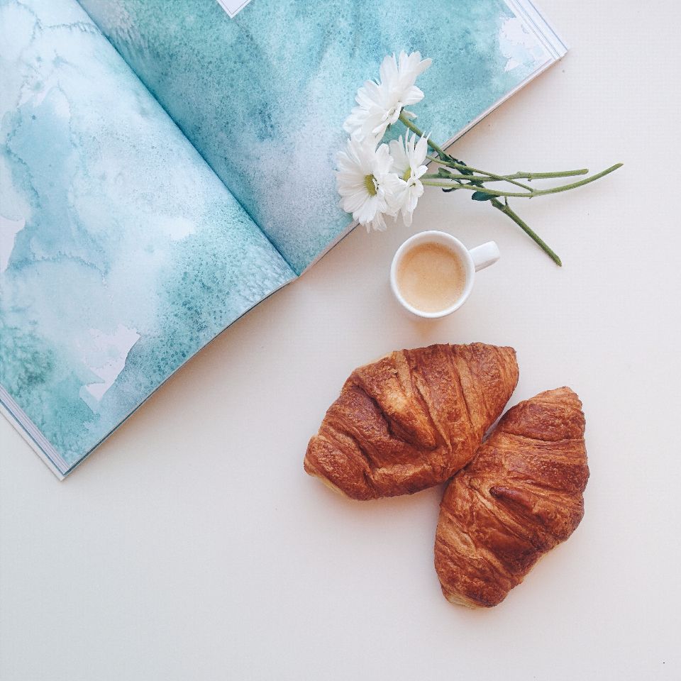 two croissants sit on a white surface with a cup of espresso and a book.