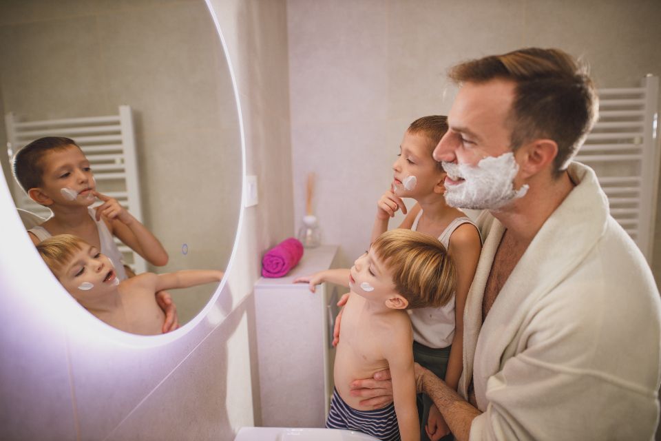 a man stands in the bathroom mirror and teaches his two young sons to shave.