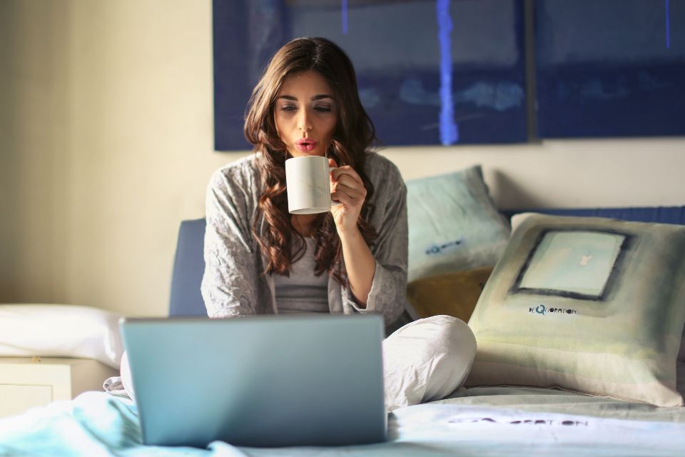 a woman sits in bed looking at her laptop and drinking from a white mug full of coffee.
